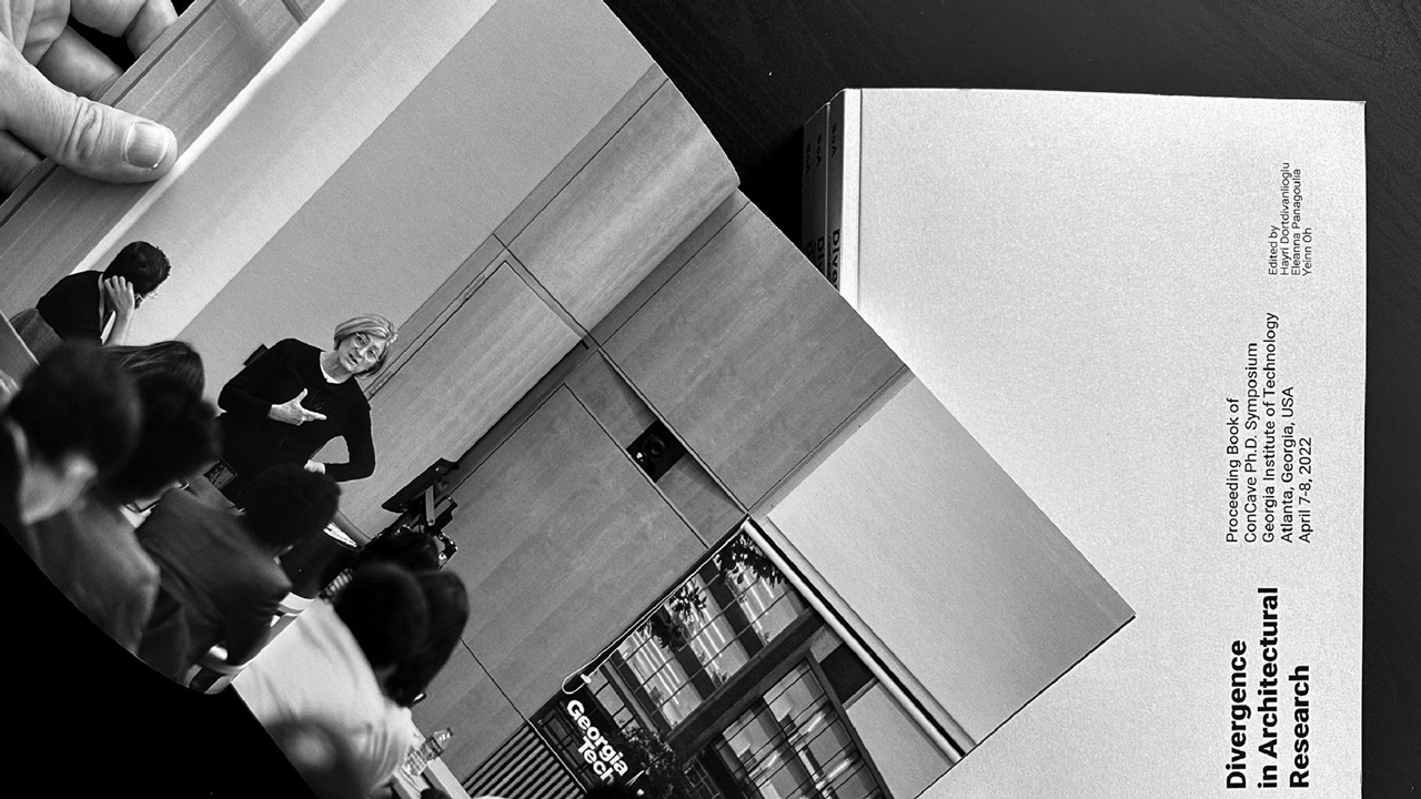 Black-and-white photo of a book being held open by a hand. The book is open to a double-page photo of Ph.D. students listening to a person speaking. Next to the open book is another copy, closed. The cover reads, "Divergence in Architectural Research; Proceeding Book of ConCave Ph.D. Symposium, Georgia Institute of Technology, Atlanta, Georgia, USA April 7–8, 2022. Edited by Hayri Dortdivanlioglu, Eleanna Panagoulia, Yeinn Oh."